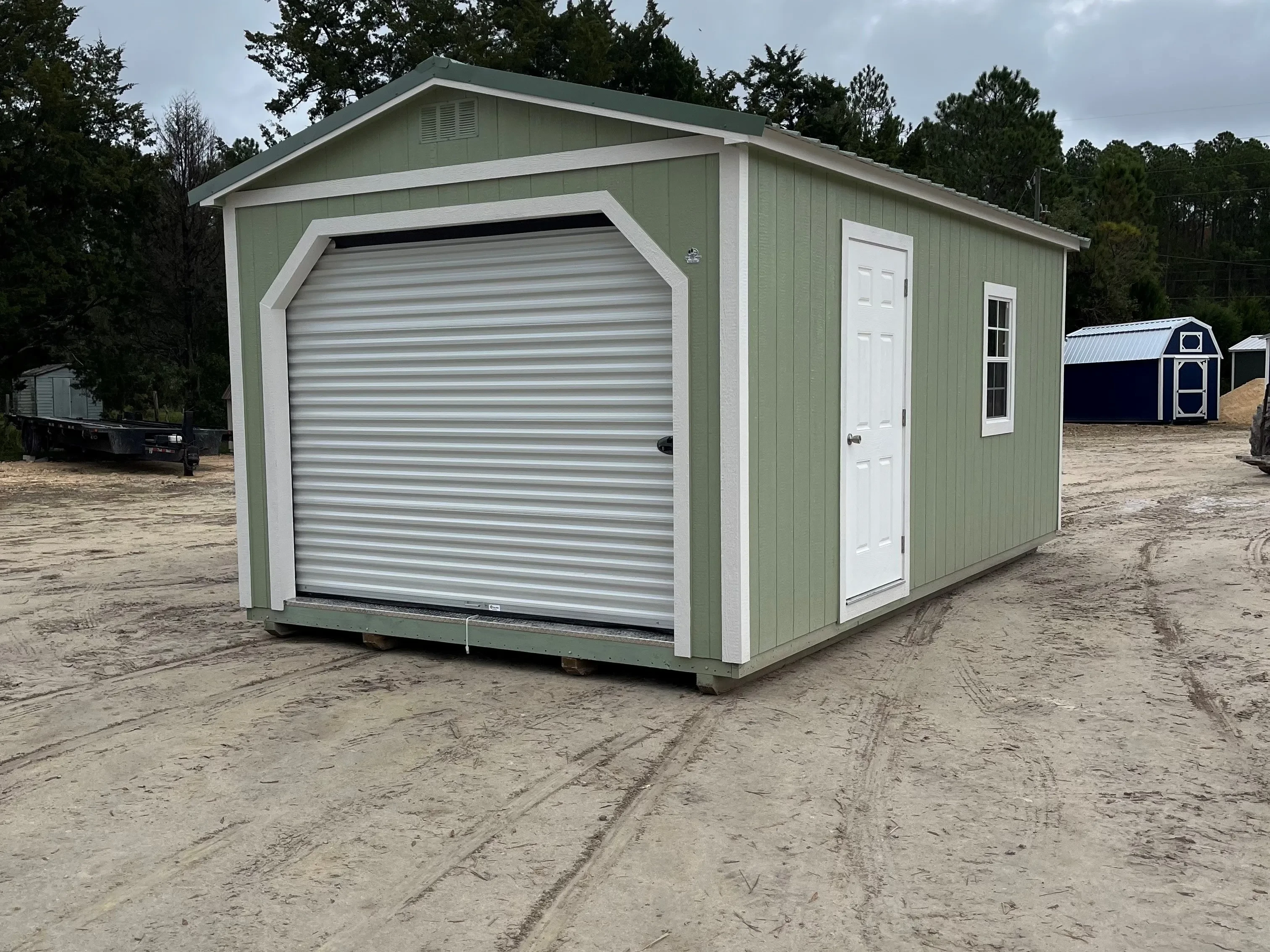 Exterior of a sage green 12x20 workshop shed showing a garage door in the end, and a walk-in door and window on the side