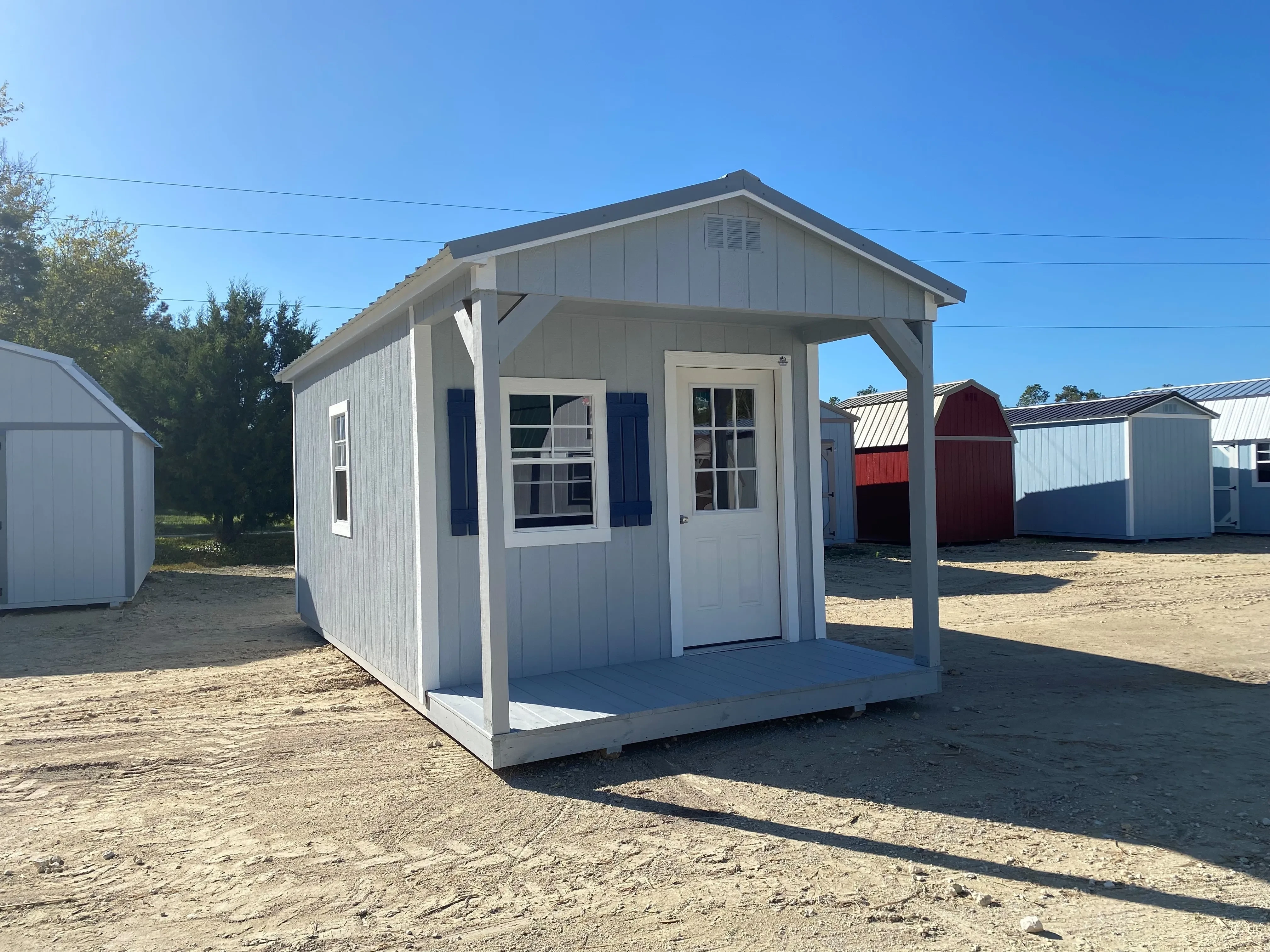 Outside of a light grey 10x20 cabin with single entry door, windows, a porch, and dark blue shutters
