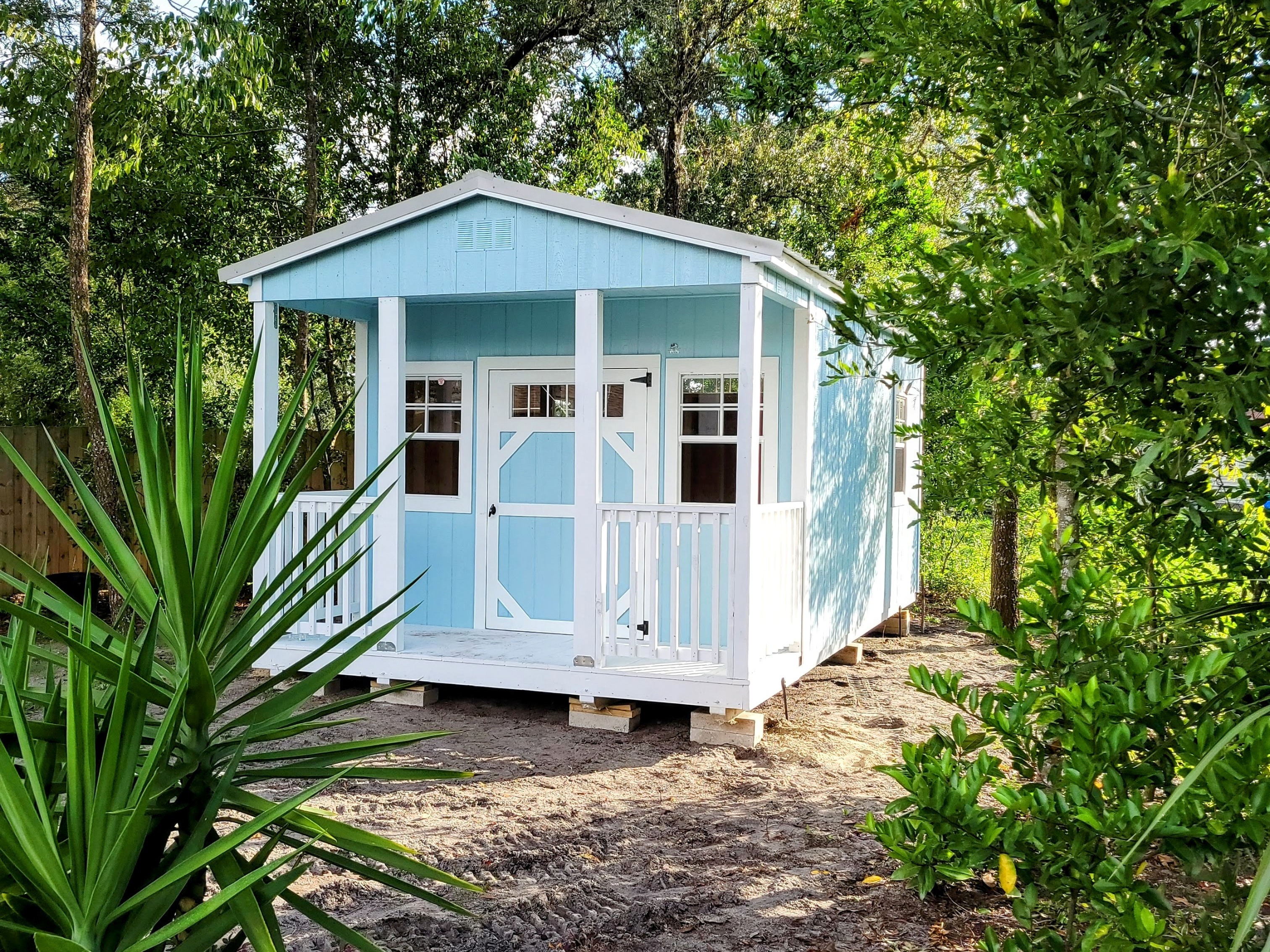 Cute light blue shed with white trim and a white porch among green Florida foliage